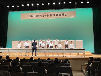 南三陸町小・中学校音楽祭の幕が飾れた舞台上に机が設置され生徒たちが2列に並んでいる写真