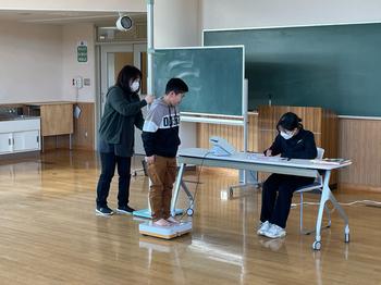 男の子が身長計と体重計の上に立ち一人の先生が背後から測定を行っており、もう一人は机に座って記録をつけている身体測定の写真