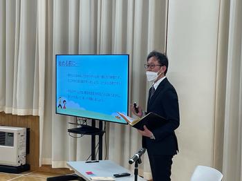 資料が映し出され大きなモニターの隣に立ったスーツ姿の男性が、手にマイクとノートを持ちながら話をしている様子の写真