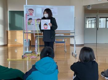 先生がホワイトボードの前で顔の表情を描いたイラストを使って説明し床に座った数人の児童が先生の話を真剣に聞いている様子を写した写真