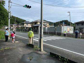 横断歩道の向こうにはお巡りさんが立ち、渡る前の女の子と2名の見守り隊の方がいる登校の様子を写した写真