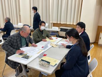 会議室で4人の参加者が資料を広げて机を囲み、書類に目を通したり意見を交わしたりしている様子を写した写真