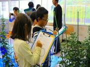 教室の中で複数の子供たちが植物を観察しながら、それぞれクリップボードにメモを書いている様子の写真