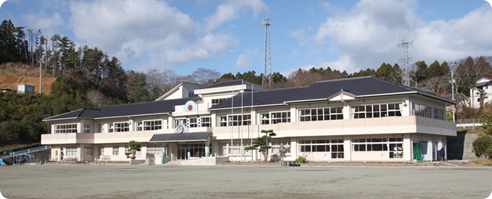 白とベージュを基調とした壁にグレーの屋根の二階建て校舎が広い砂のグラウンド前に立っている名足小学校の外観写真