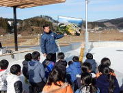 屋外で男性がパネルの写真を持ち上げ複数の子供たちに向かって見せながら説明している写真