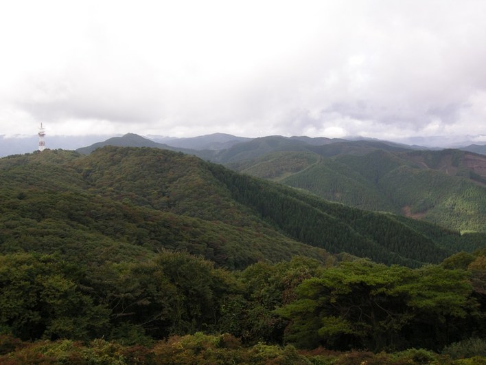 分厚い雲がかかる空の下、山の尾根に電波塔が立つ深い緑の山並みの写真