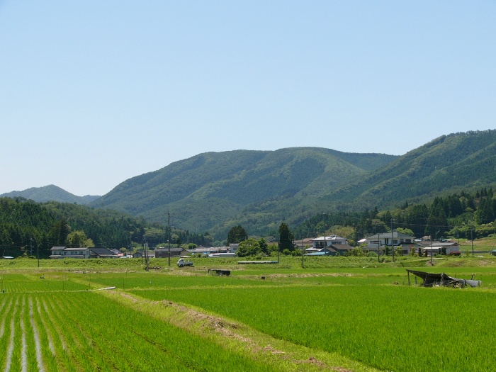 青空の下、田んぼと民家が広がり、奥に穏やかな山並みが連なる風景写真