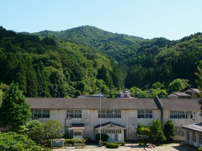 山あいの小学校の校舎が中央にあり、背景に新緑の山が広がる風景の写真