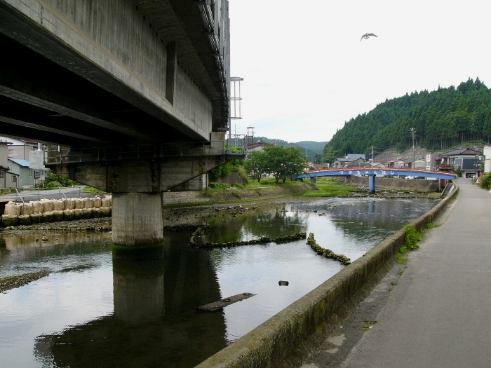手前に大きなコンクリート橋の橋脚が立つ川沿いの景色で、奥には赤い橋と緑の山々が見える写真