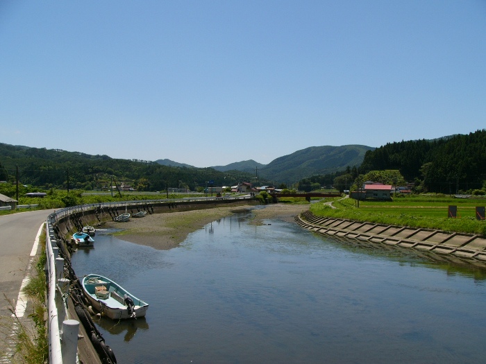 青空の下、水位の低い川に小型ボートが浮かび、両岸に道路と田畑が広がる風景写真