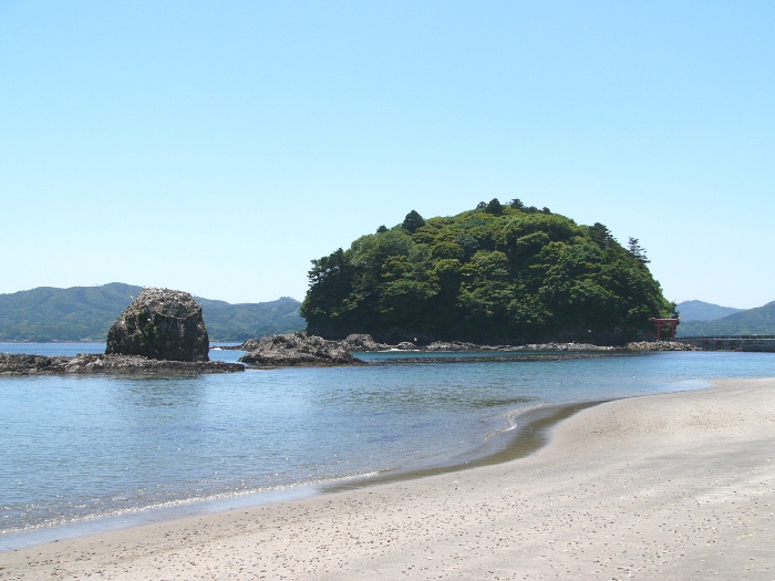 白い砂浜とその先に入り口に鳥居が立つ緑豊かな小島が浮かぶ、晴れた日の穏やかな海岸の写真
