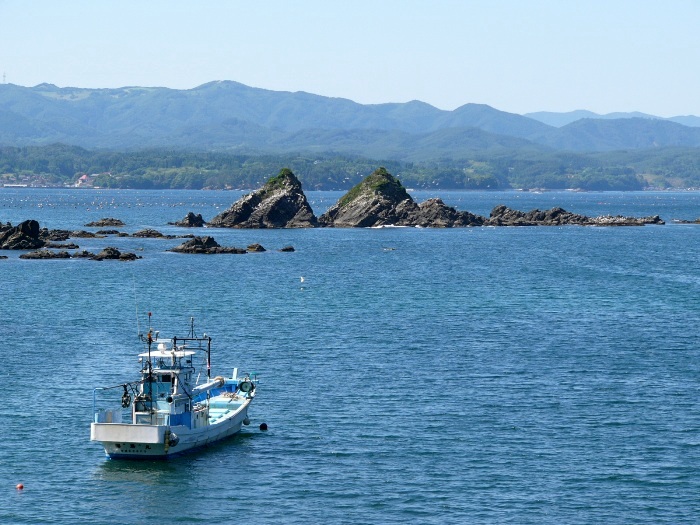 青い海に浮かぶ漁船と、奥には岩が連なる小さな島が見える写真