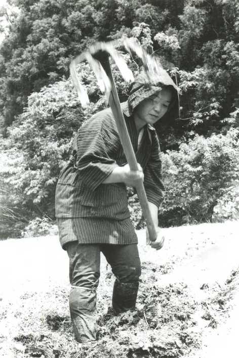 作業着を身にまとい、頭には頭巾ようなものをかぶった女性が手に大きなクワを持ち、土を耕している様子の白黒写真