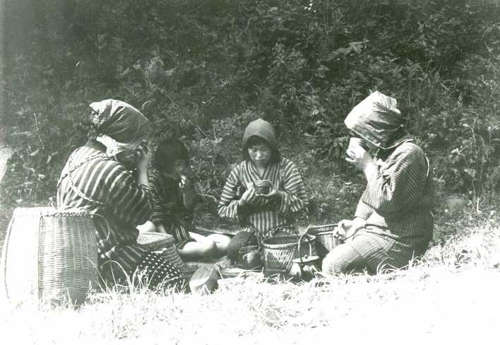 作業着と布をかぶった3人が草の上に囲んで座り、休憩をしている様子の白黒写真