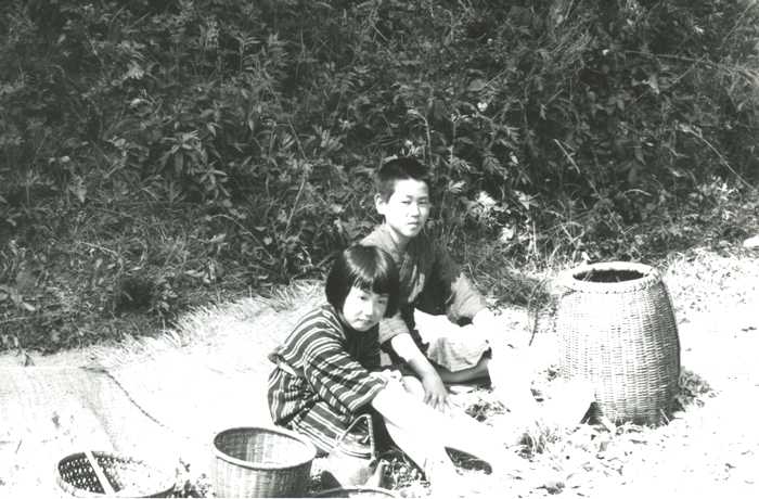 竹かごなどが置かれたそばで女の子と男の子がござの上に座っている様子の白黒写真