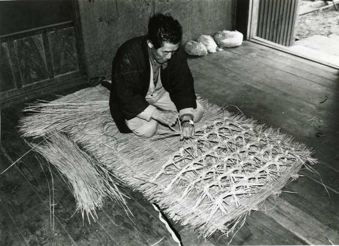 室内で、男性が床に座り手で藁を編んでいく作業中の様子を撮影した白黒写真