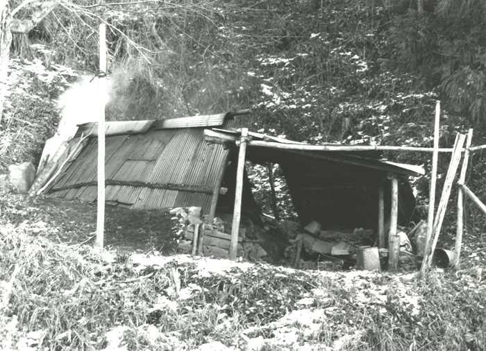 山の斜面に建っている木や竹を使って作られた、小さな小屋から白い煙が立ちのぼっている炭焼き釜を正面から撮影した白黒写真