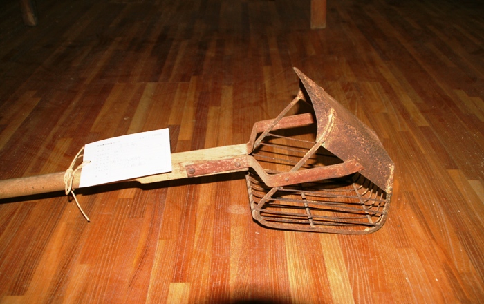 木の柄の先端に鉄製のかご状の器具がついた土入れ機の写真