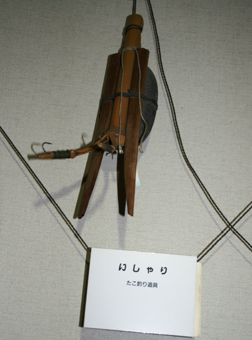 「いしゃり」と書かれた札の上に、筒状の竹と2本の木と鉄でできたタコ漁専用の漁具が壁に展示された写真