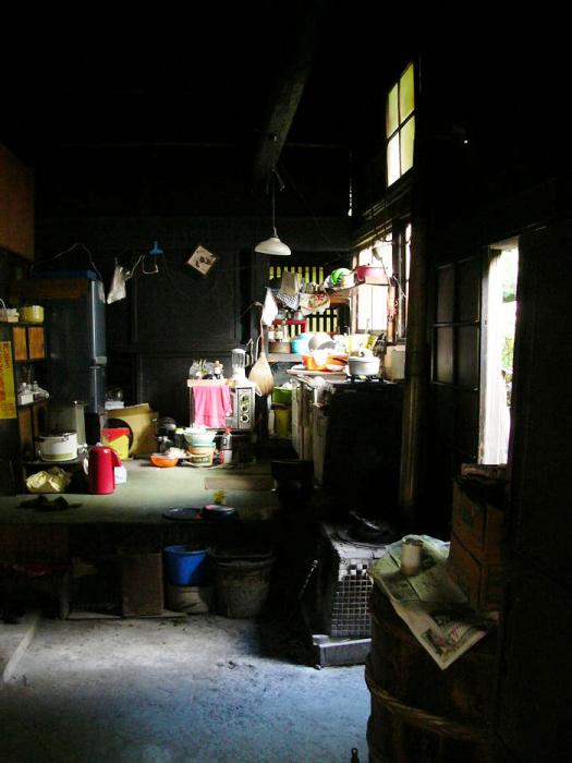 窓から太陽光が差し込んだ薄暗い土間にガスコンロや冷蔵庫などの台所道具が置かれた昔の炊事場の写真