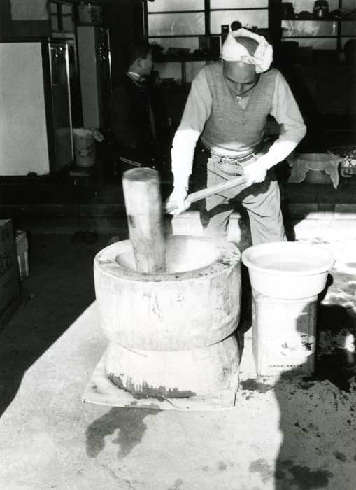 頭にタオルを巻いた男性が大きな臼で杵を使って餅をついている白黒写真