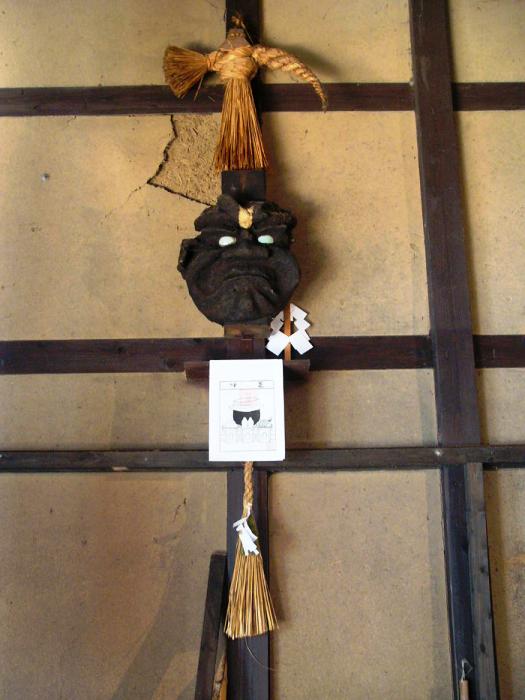 土壁と木枠の家屋内に、藁の飾りや紙垂とともにカマドの神ともいわれる「かま神さま」の黒い面が祀られている写真