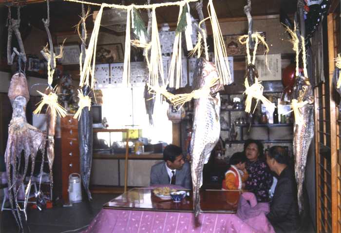 家族がこたつを囲んで談笑している居間にしめ縄や飾り、魚やイカ、タコが吊るされている正月のかけざかなの様子を写した写真