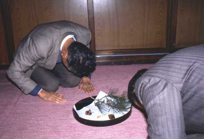 三角形の餅2個・榧の実・串柿（干し柿）を載せ、松の葉を添えた小型の盆を挟んで、正座した2人の男性が頭を下げて挨拶をしている様子の写真