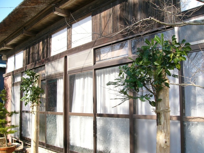 日本の古い家屋の縁側前に、両脇に蔦や緑の葉をつけた枝が束ねられた木の柱が立てられている写真