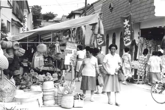 露店が並ぶ賑やかな通りで、買い物を楽しむ人々や竹籠などの生活用品が売られている盆市の様子を写した白黒写真