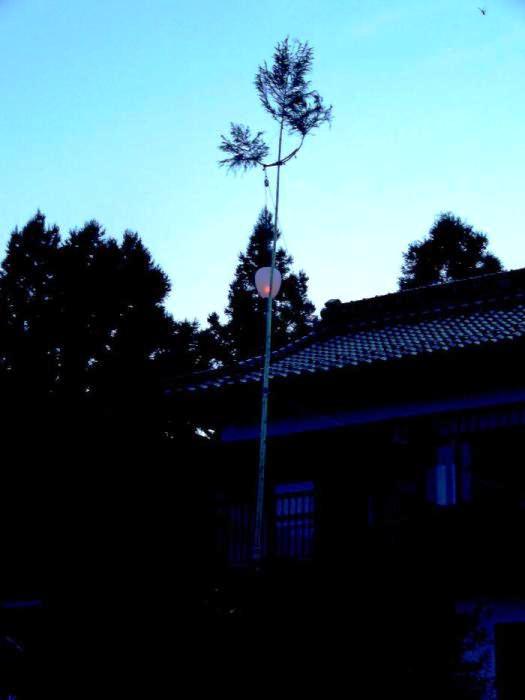 古民家の前に梢を残して樹皮を剥いだ若杉の柱が立てられ、この柱に灯籠が取り付けられ夕暮れの空に浮かび上がっている灯篭柱を写した写真