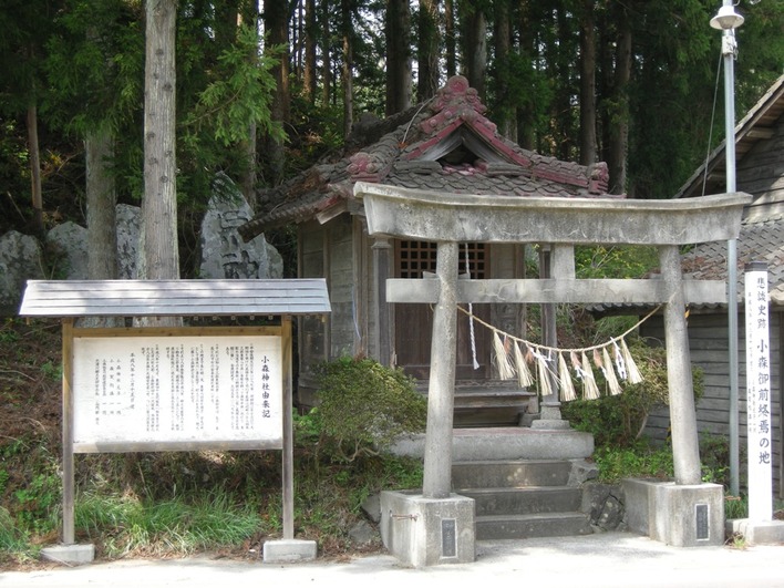 木造の小さな社殿の前に注連縄のかかった石造りの鳥居があり、左には由来を記した案内板が立ち、背後には石仏が並び杉林が広がっている小森御前社（志津川小森）の写真