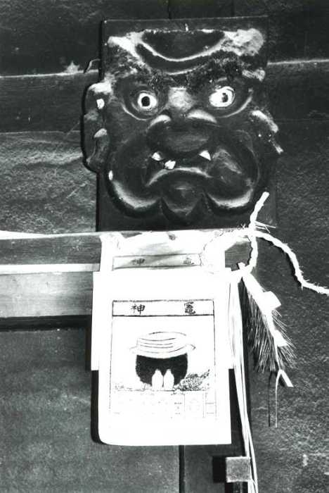 鬼のような顔をした木彫りの面のかま神さまが梁に取り付けられている白黒写真