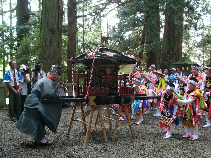 森の中の神社境内で神輿を前に伝統衣装をまとった子どもたちが並び、神職が儀式を行っている様子が写っている写真