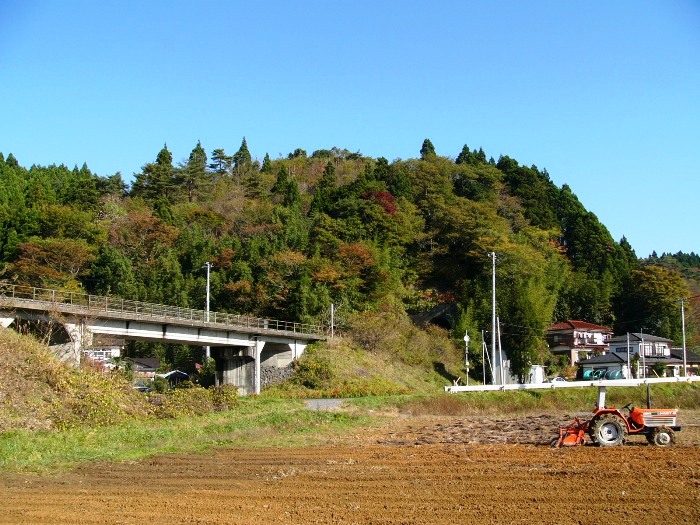 手前に耕された畑、左手には高架道路、奥には紅葉しだした緑濃い山がある要害館跡の写真