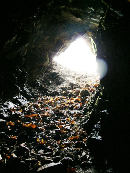 岩肌に囲まれた細い坑道の内部から外を見た様子で、地面には落ち葉や小石が散らばり、奥の出口が白く強い光で照らされている十枚山産金跡の写真