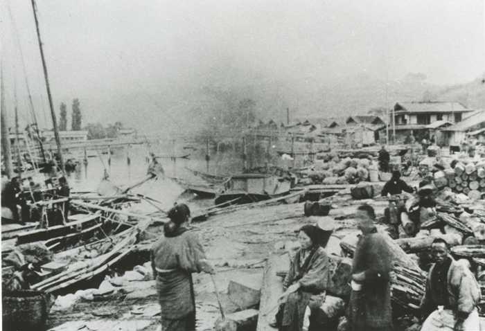 川沿いの港に複数の船が停泊しており、岸には木材や荷物が大量に積まれていて、物流や作業の拠点として活用されている様子が写された古い写真