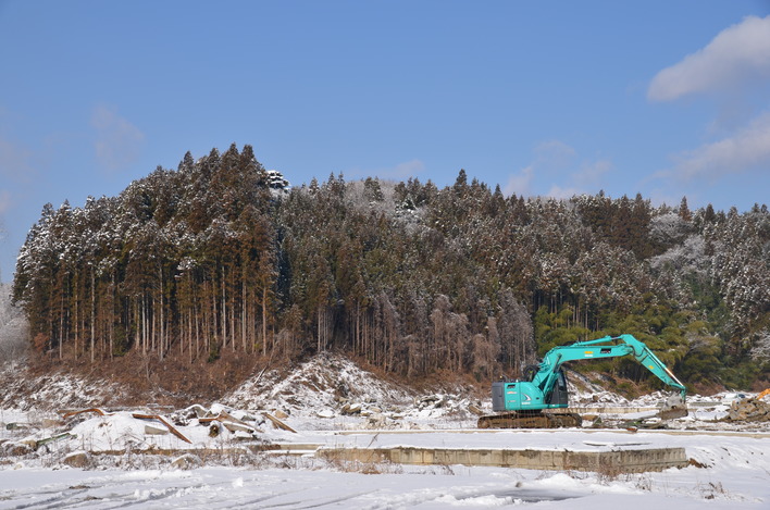 雪に覆われた地面と建設跡地の前に緑色の大型ショベルカーがあり、背景には雪化粧した杉林の小高い山が広がる新井田館跡の写真