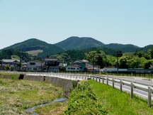川と道路が並んで走り、その先に丸みを帯びた山々が見える長閑な町の写真