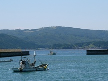 防波堤の間を漁船が進み、奥に広がる山と海が見える港の写真