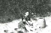 竹かごなどが置かれたそばで女の子と男の子がござの上に座っている様子の白黒写真