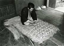 室内で、男性が床に座り手で藁を編んでいく作業中の様子を撮影した白黒写真