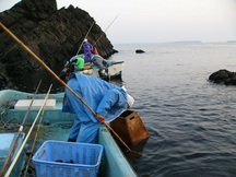 カッパを着た漁師が、岩場の近くに小型の漁船を停めて海に網や仕掛けを下ろしたり、箱メガネで海底を覗きこんでいる様子の写真