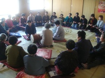 和室に大勢の人々が1本の長い縄のようなものを持ち、中央の太鼓を囲んで円になって座り念仏講が行われている写真