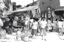 露店が並ぶ賑やかな通りで、買い物を楽しむ人々や竹籠などの生活用品が売られている盆市の様子を写した白黒写真
