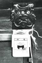鬼のような顔をした木彫りの面のかま神さまが梁に取り付けられている白黒写真