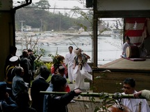 海辺の神社で伝統的な衣装を着た人物が舞を演じ、周囲には竹を手にした人々や太鼓を叩く演奏者が集まり、例祭が行われている様子が写っている写真