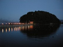 夕暮れ時に海に浮かぶような小島へ続く橋が灯籠の明かりで照らされ、鳥居が島の入口に立っている神秘的な風景が写っている写真
