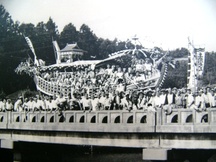 橋の上に大勢の人々と共に巨大な龍の装飾を施した船型の山車が乗っており、祭りの一場面と思われる活気ある様子が写っている白黒写真