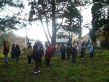 背の高い木々に囲まれた林内の広場で、看板前に集まり説明を聞いている人々の様子を撮影した羽黒山清水寺跡の写真